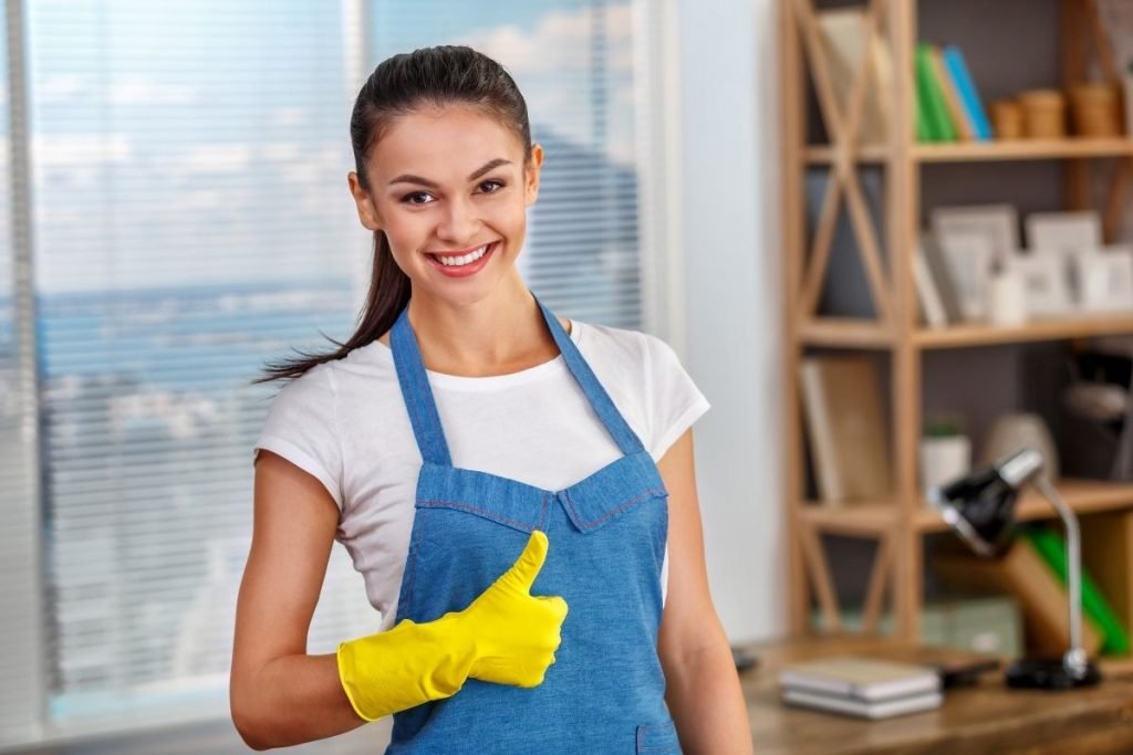 A woman in an apron and rubber gloves holds a yellow glove, preparing for cleaning or cooking tasks.